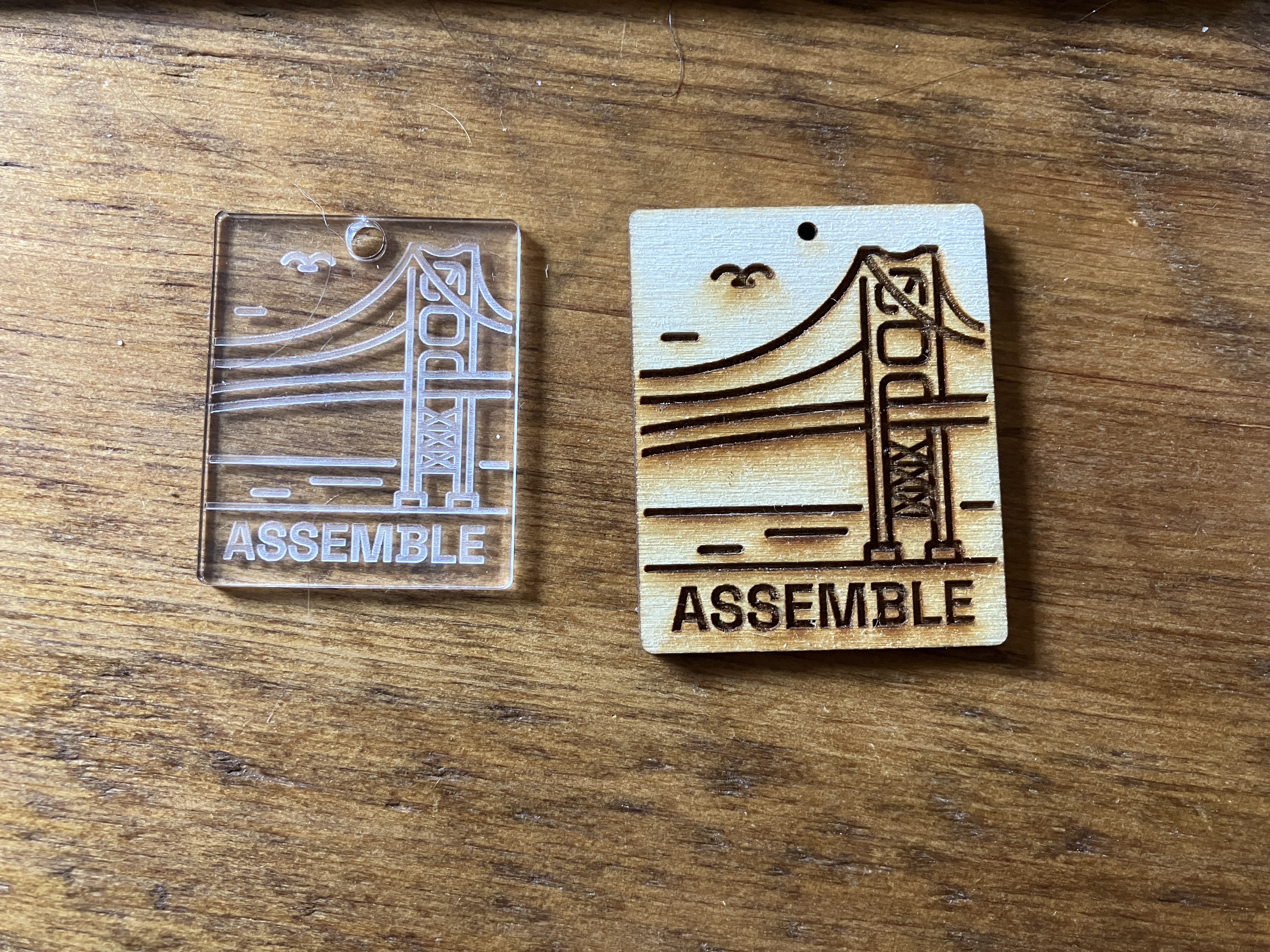 Two rectangular pieces laying flat on a wooden table engraved with the Assemble logo. The left piece is made with transparent plastic, and the piece on the right is wood.